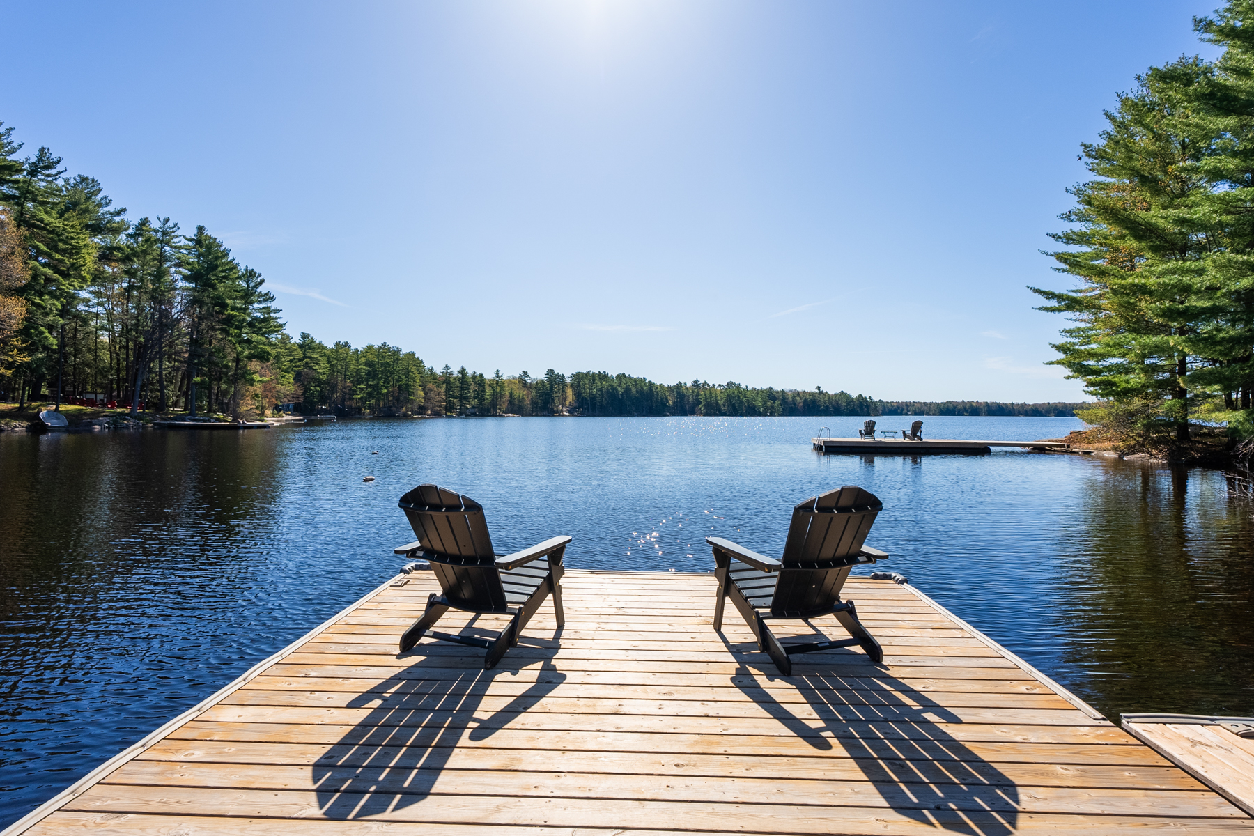 Two Muskoka chairs sit on the end of a wooden dock, looking out across a blue lake.