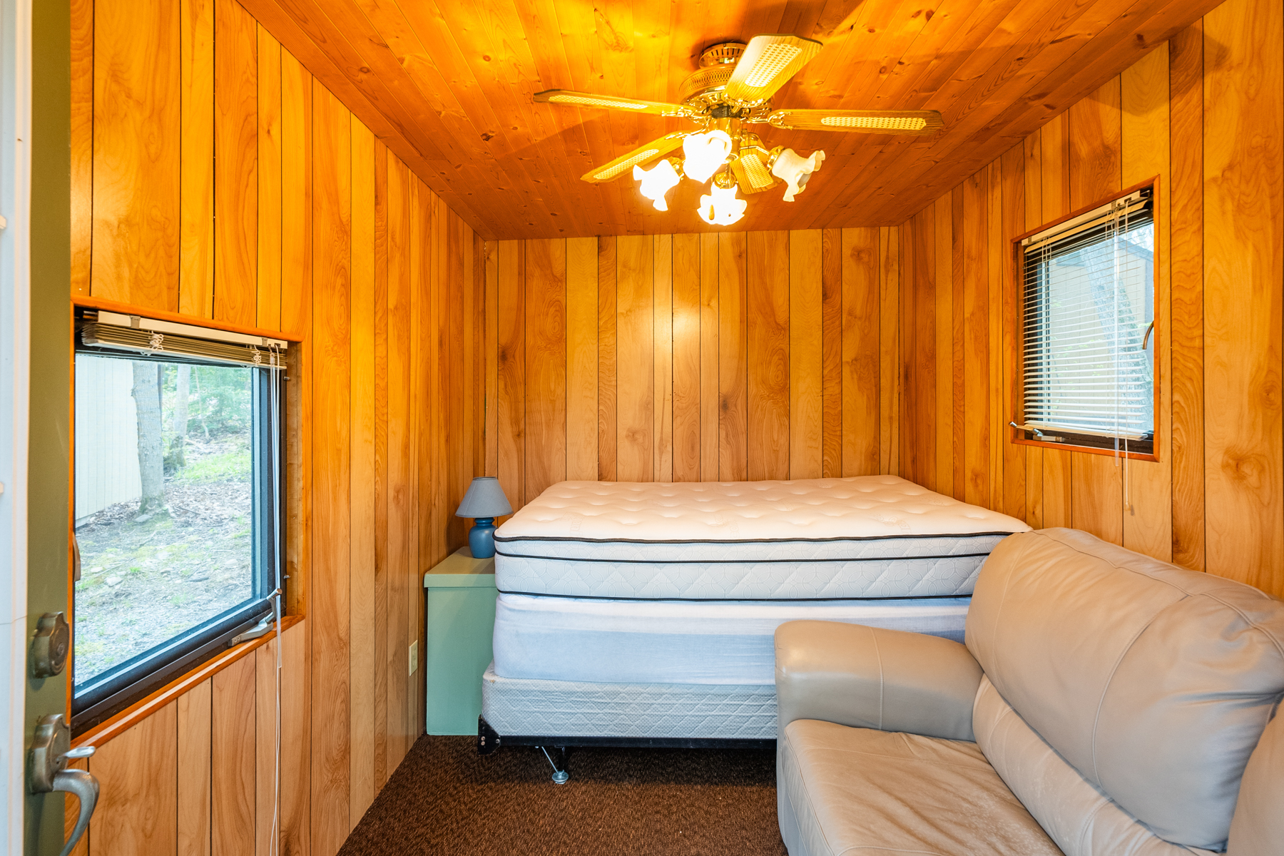 A wood-panelled interior of a small cabin with a couch, a ceiling fan, a mattress, and two windows.