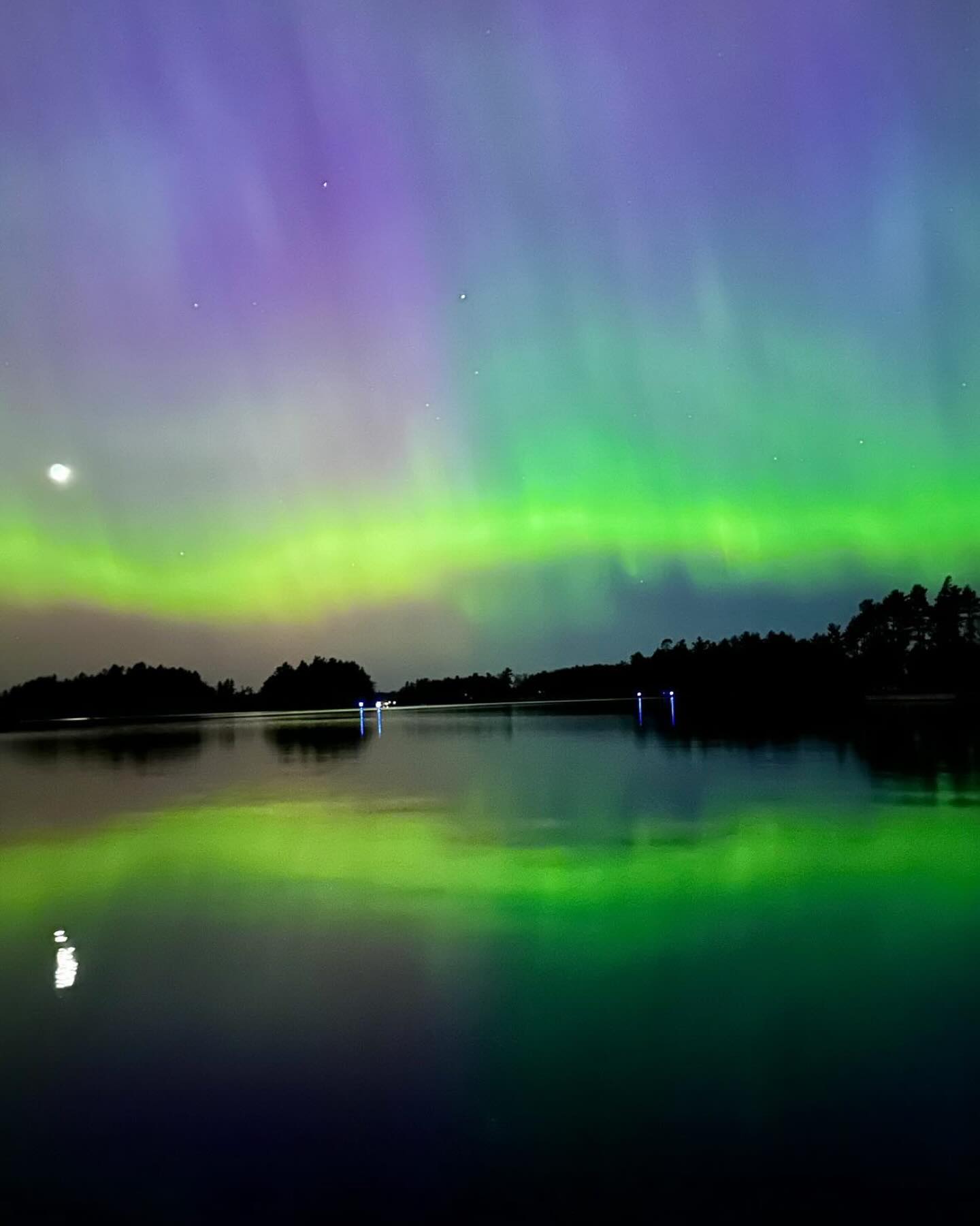 Northern lights appear over a still lake with tree line silhouette