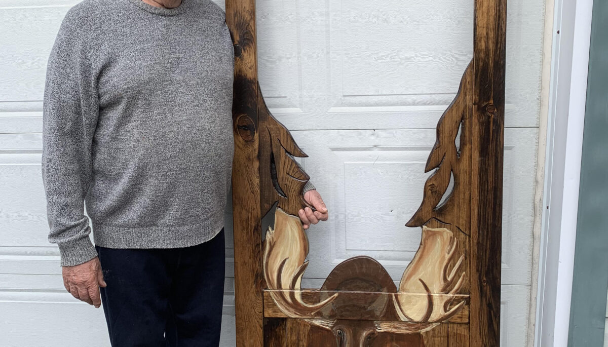 A person in a grey sweater smiles while standing beside a dark brown screen door with a moose painted on the bottom