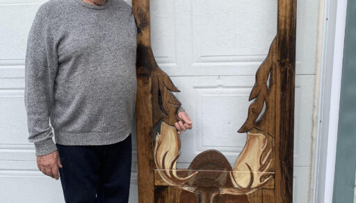 A person in a grey sweater smiles while standing beside a dark brown screen door with a moose painted on the bottom