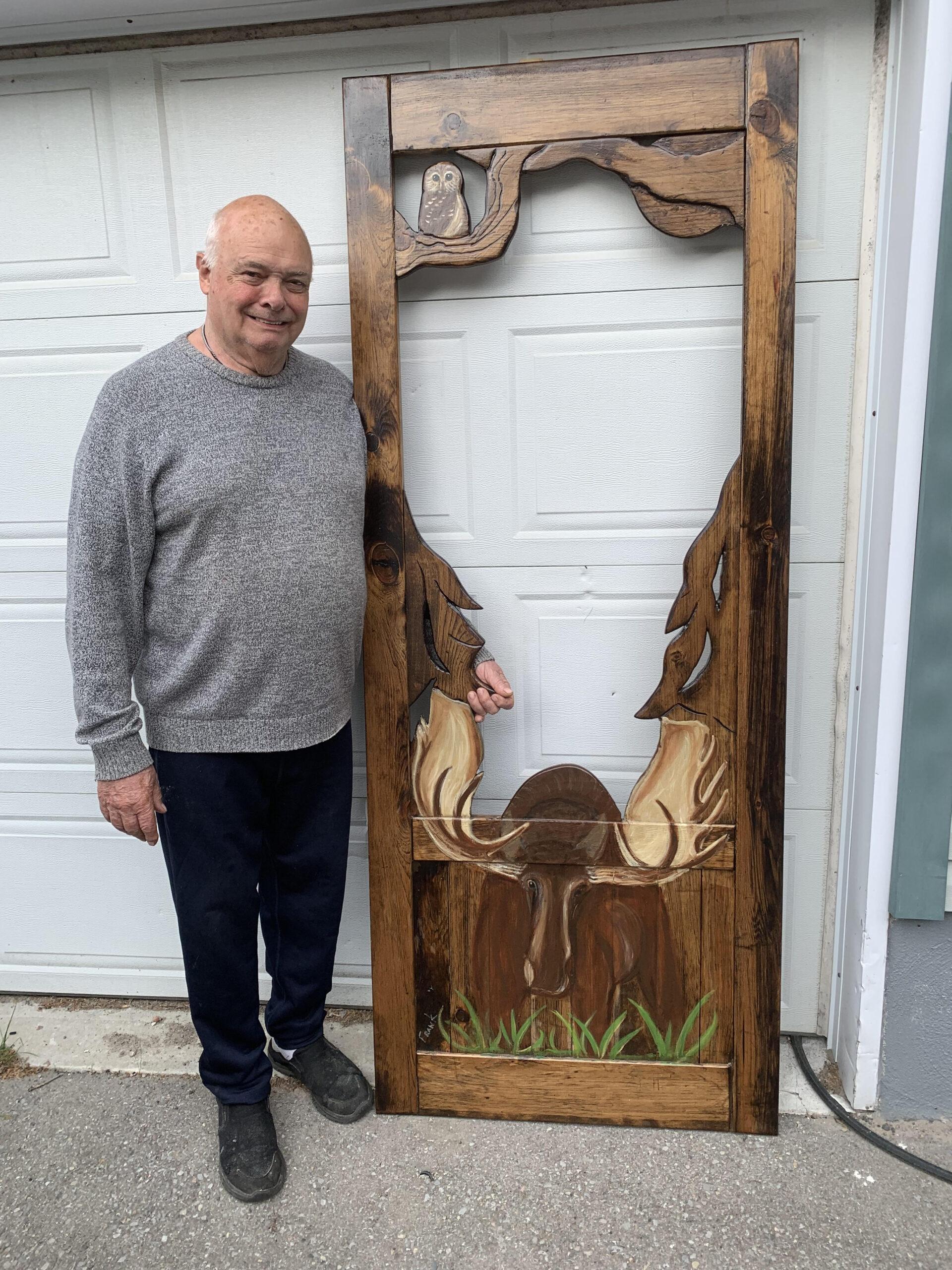 A person in a grey sweater smiles while standing beside a dark brown screen door with a moose painted on the bottom
