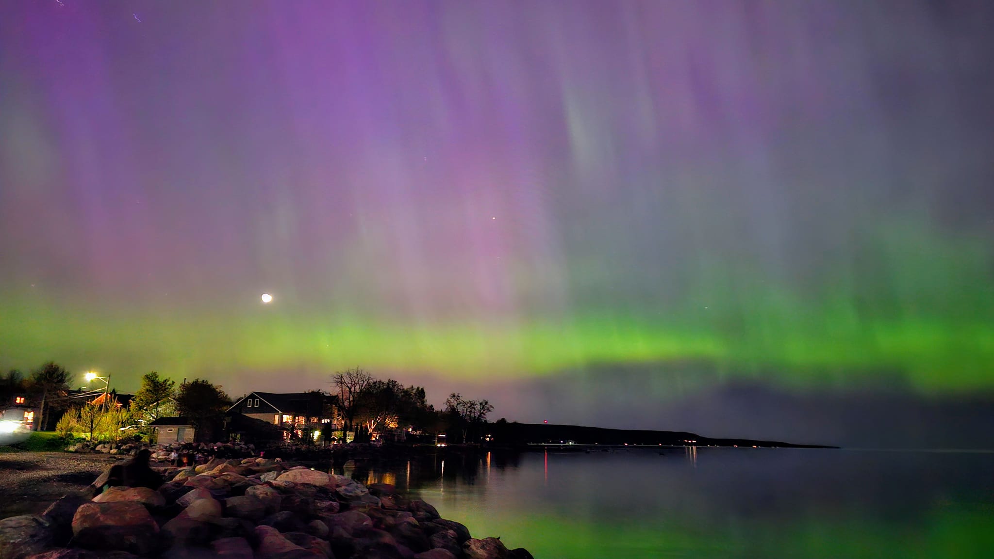Northern lights over a rocky shoreline with buildings in the distance