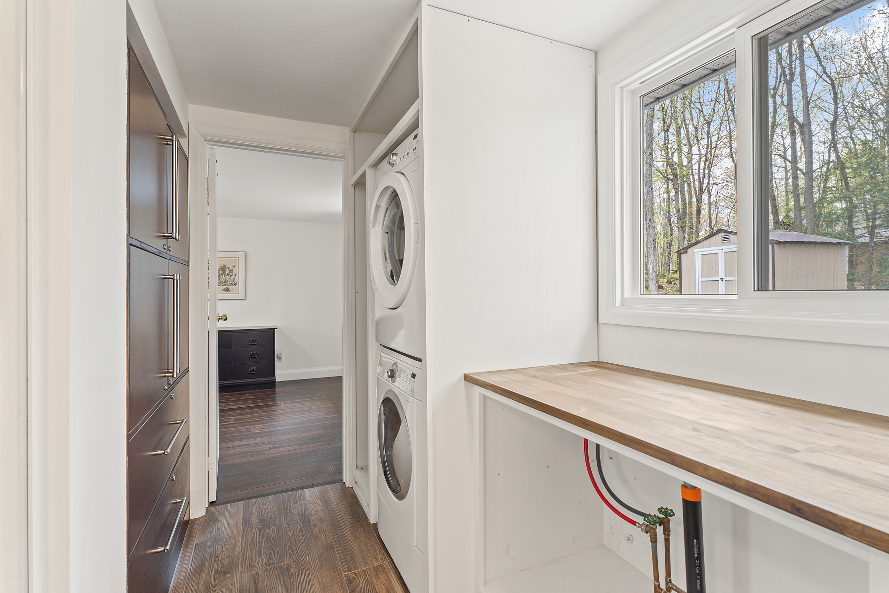 A laundry area in a bright, narrow hallway, with a stacked washing machine and dryer set into a closet flush with the wall, across from a wall of storage cupboards and drawers.
