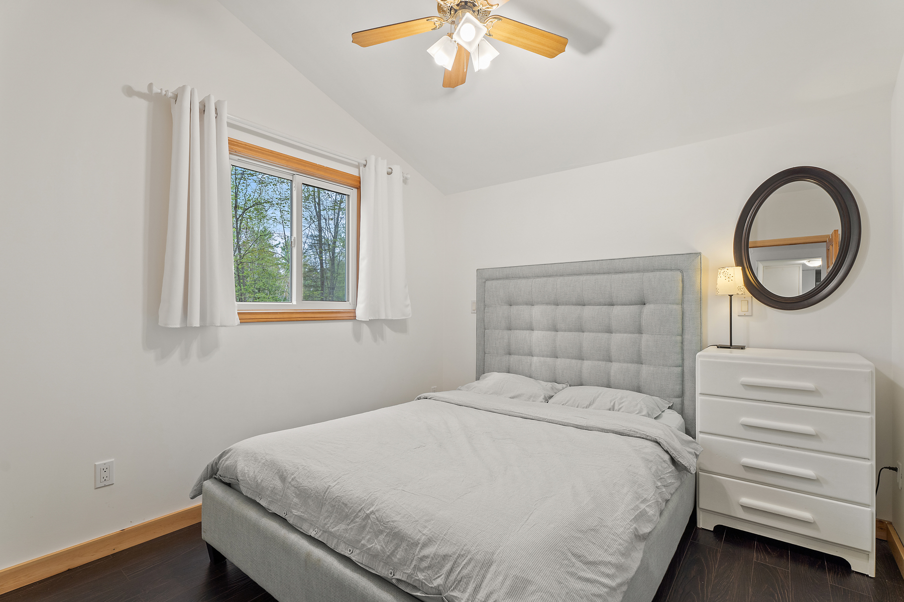 A bright, simple bedroom with hardwood flooring, a ceiling fan, a window, and a small dresser.
