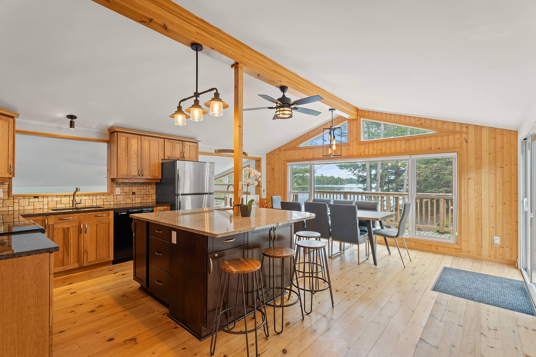 A large, open-concept kitchen with a dining area, which has a big table and chairs, and large windows across a wood-panelled accent wall with a vaulted ceiling.