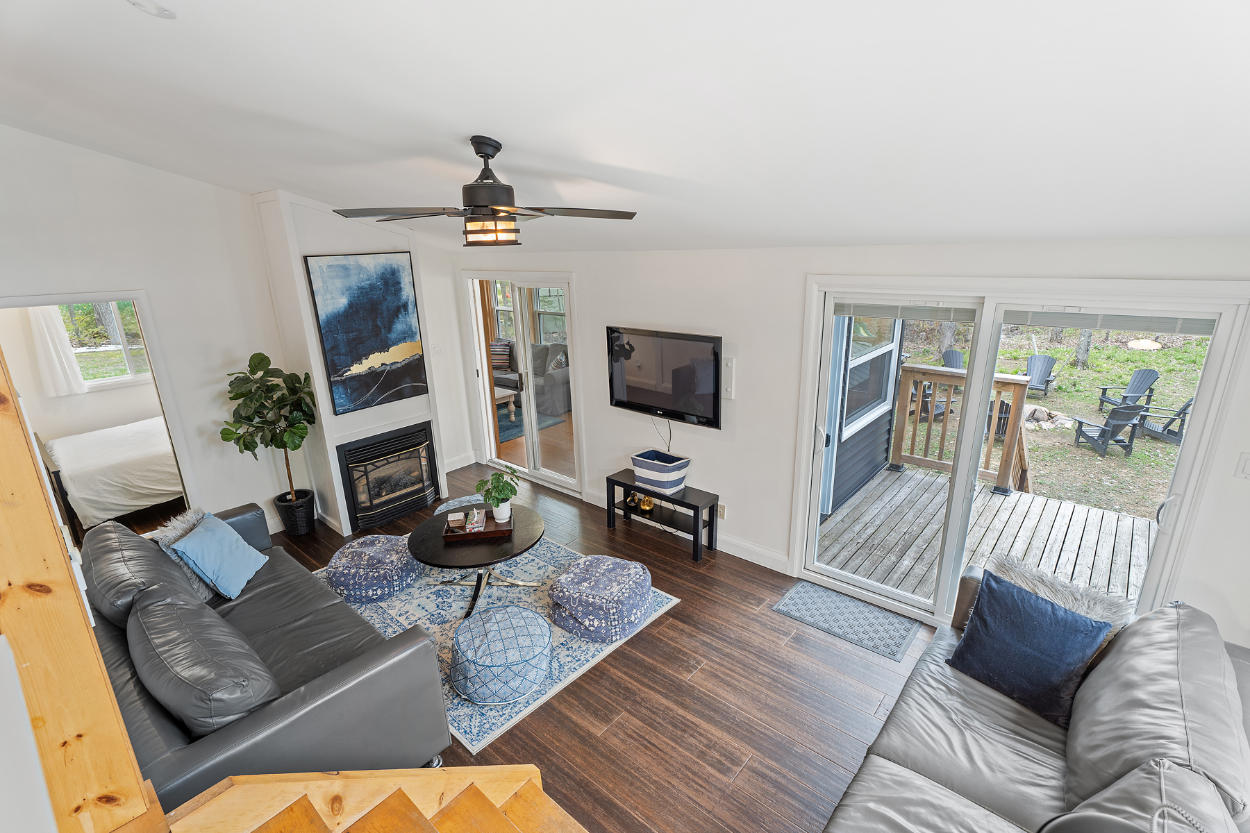 Overhead view of a cozy living room with two couches, a television, a propane fireplace, and high ceilings. A door on the left leads from the living room into a bedroom, and two sets of sliding glass doors on the adjacent wall lead out to a sunroom and a deck.