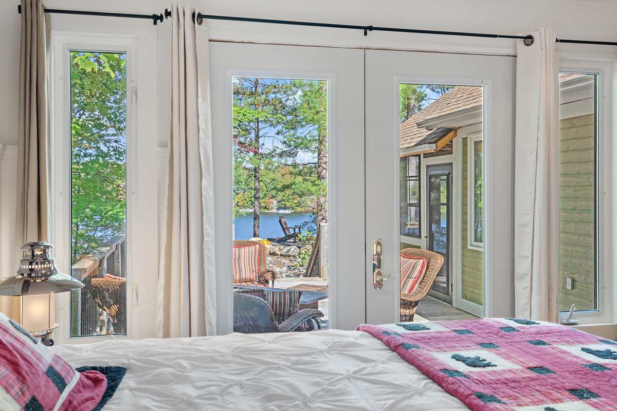 A bed is right next to bright doors with white frames that look out onto the deck