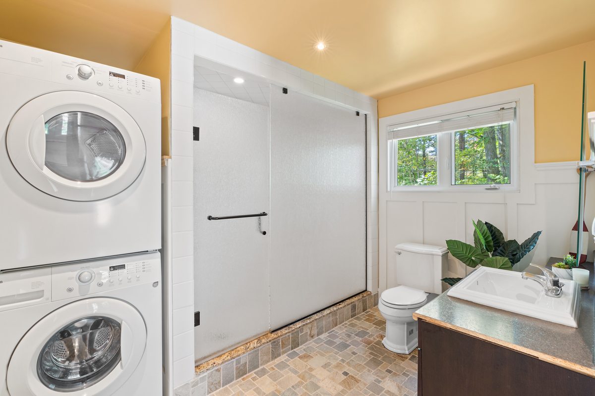 Stacked laundry machines to the left of a cloudly-glass shower, next to a toilet