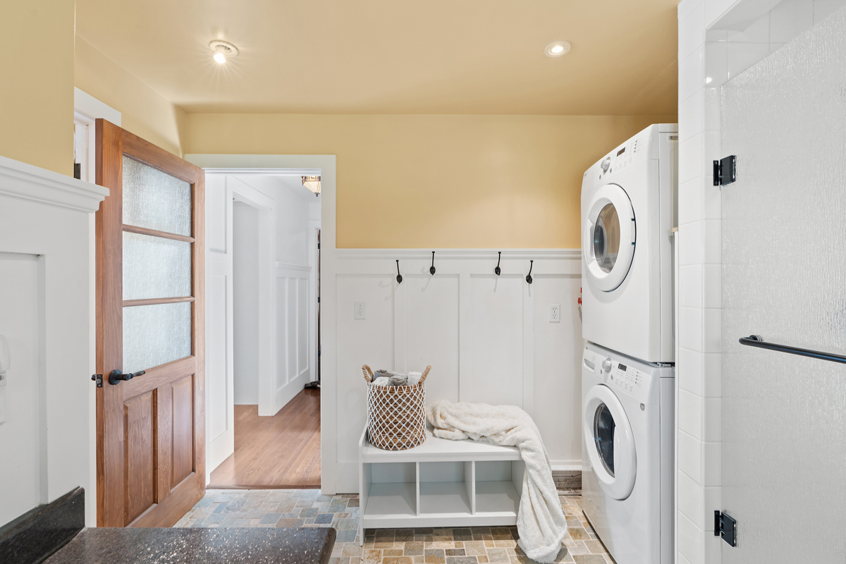 Stacked laundry machines next to a white bench with storage, and an open door leading back into the cottage