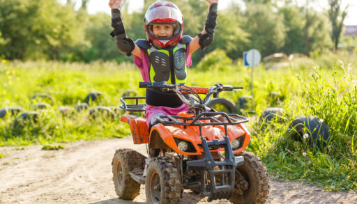 A young girl wearing a helmet and protective gear gives a double-thumbs-up while sitting on an ATV