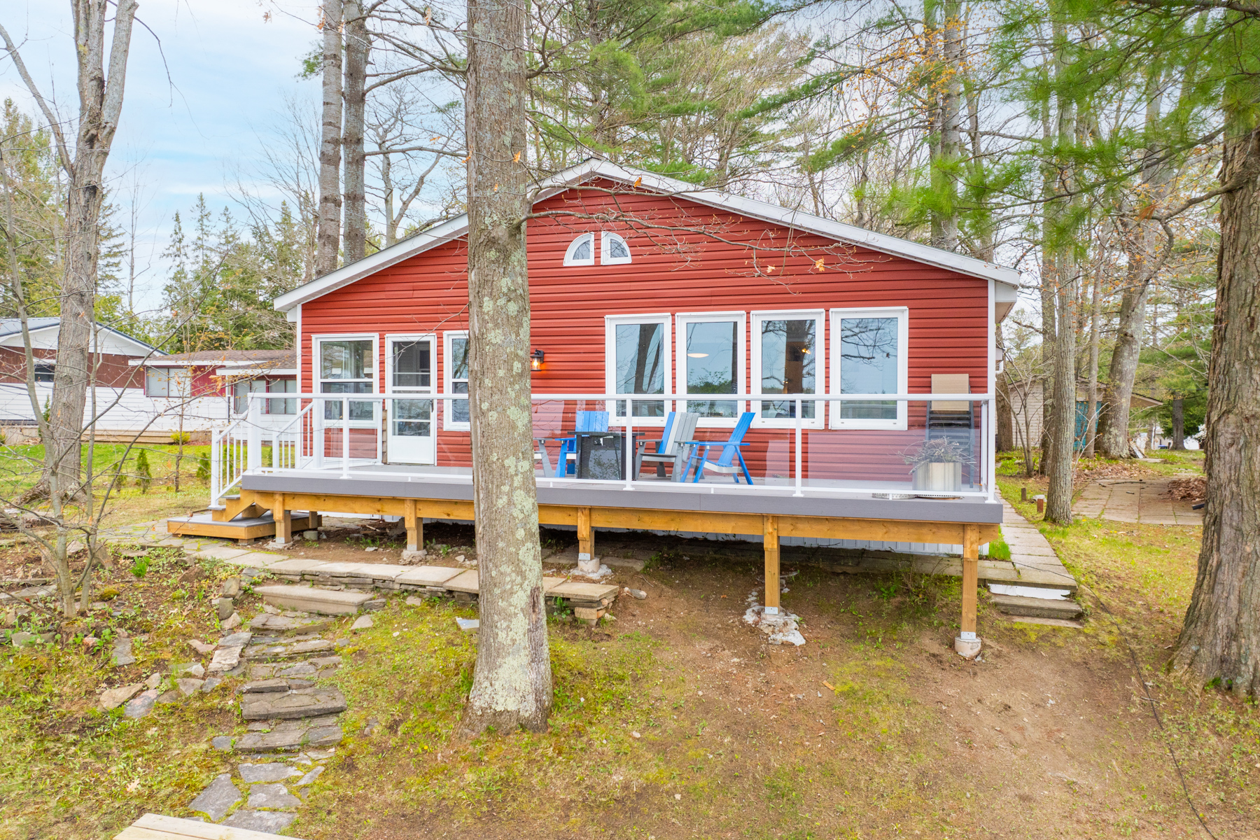 A red cottage with big windows, surrounded by a few tall trees. A new deck with a glass railing spans the length of the cottage.