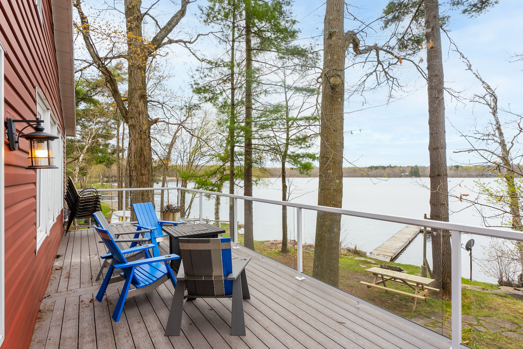 Three Muskoka chairs and a small table sit on a deck with a glass railing, looking out toward a lake.