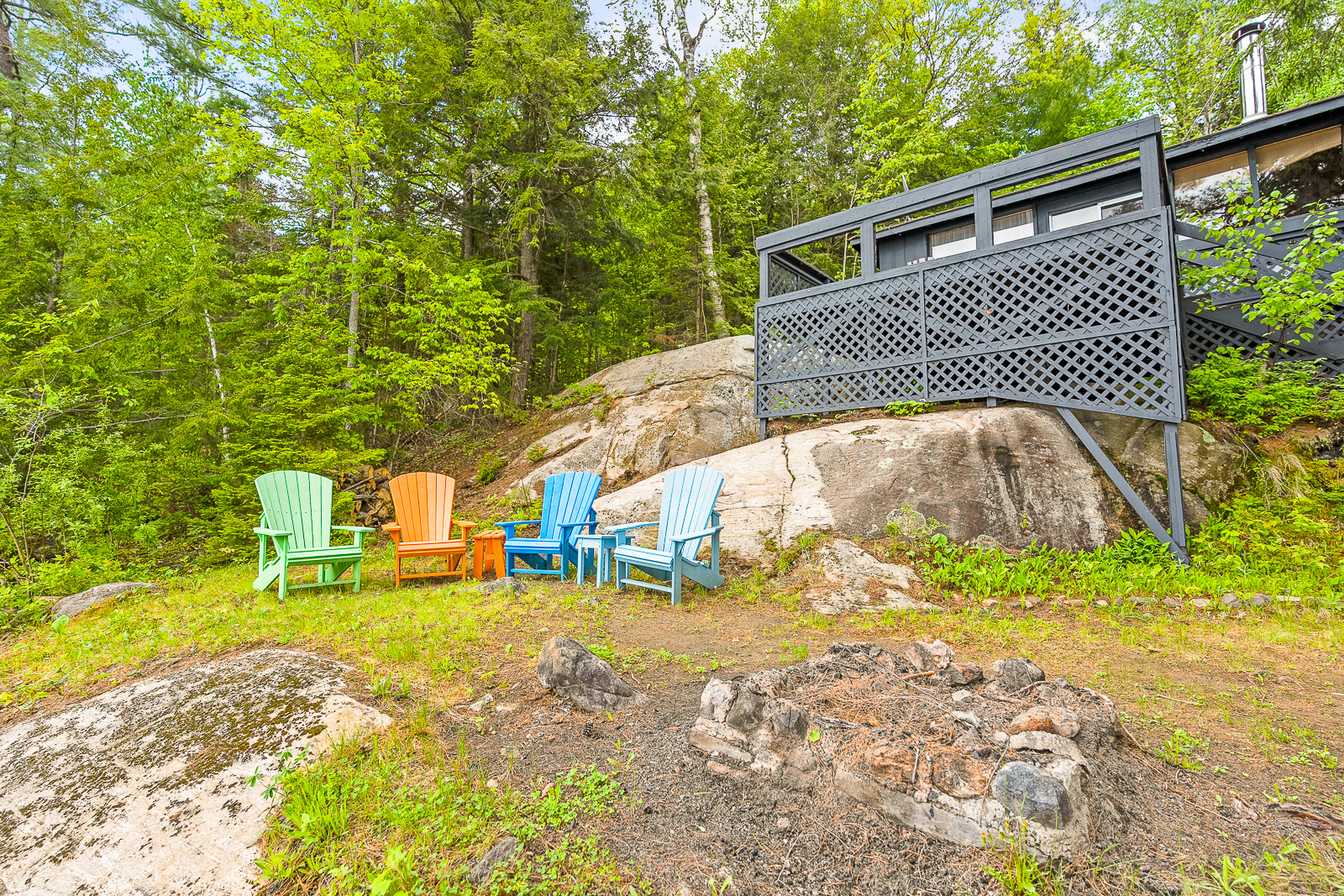 Below the grey cottage sits four coloured Muskoka chairs around a fire pit