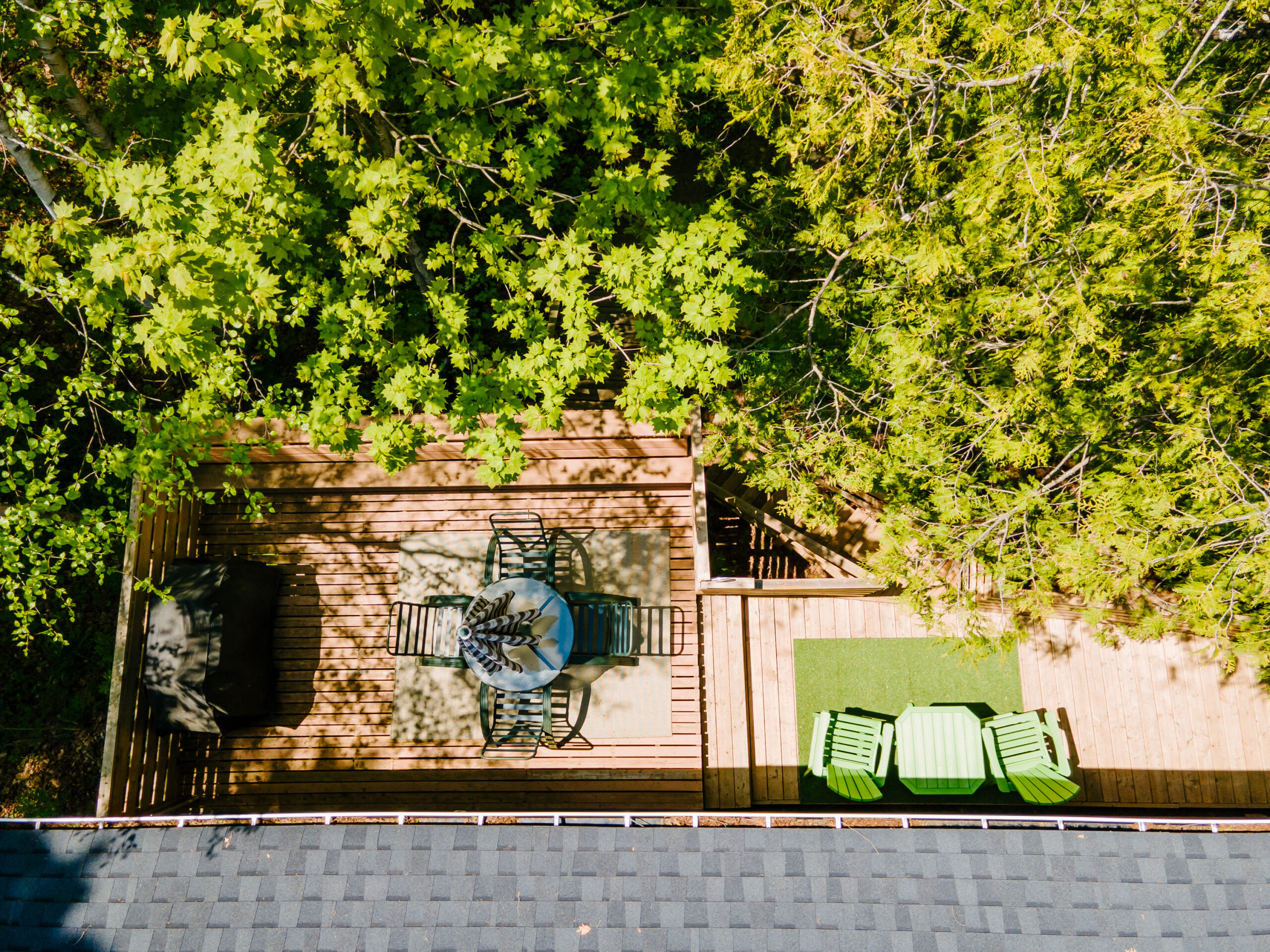 Overhead view of the back deck. From left to right: a closed striped umbrella on a glass dining table with chairs, a small fake patch of grass with green chairs and a small table