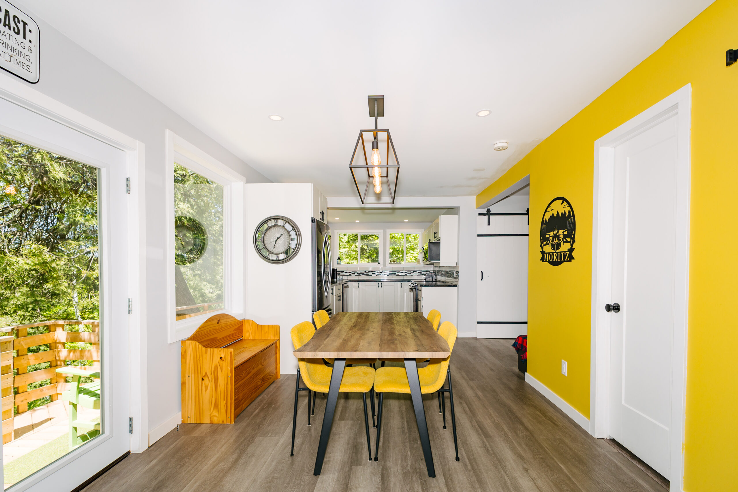 A small wood dining table with yellow chairs. White doors on the left and a yellow wall on the right. The kitchen is behind