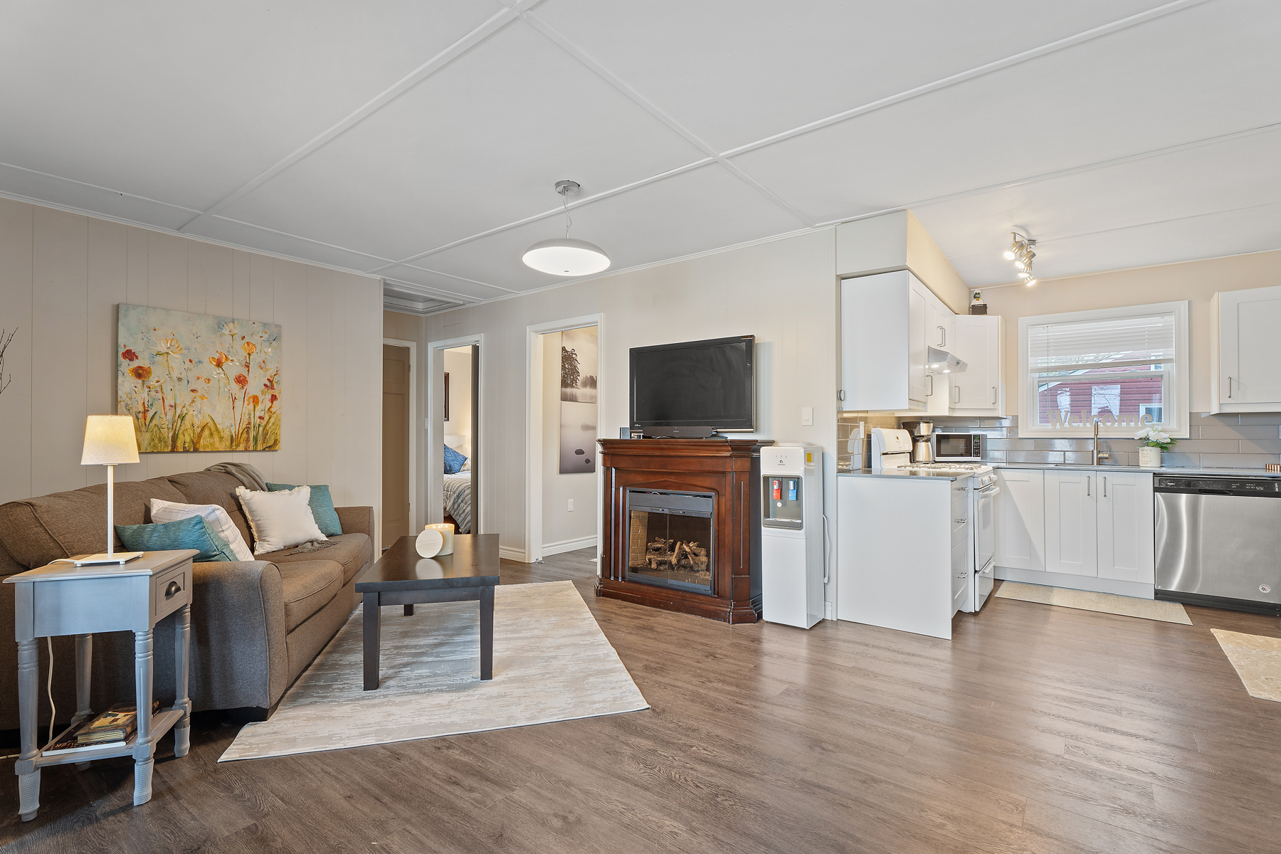 An open-concept living and kitchen area. A couch sits in front of a fireplace with a television over top. Through the living area are doorways leading to bedrooms.