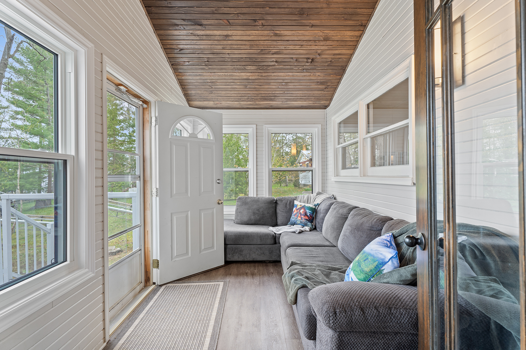 A bright sunroom with a big grey couch, lots of windows, and a wood-paneled ceiling.