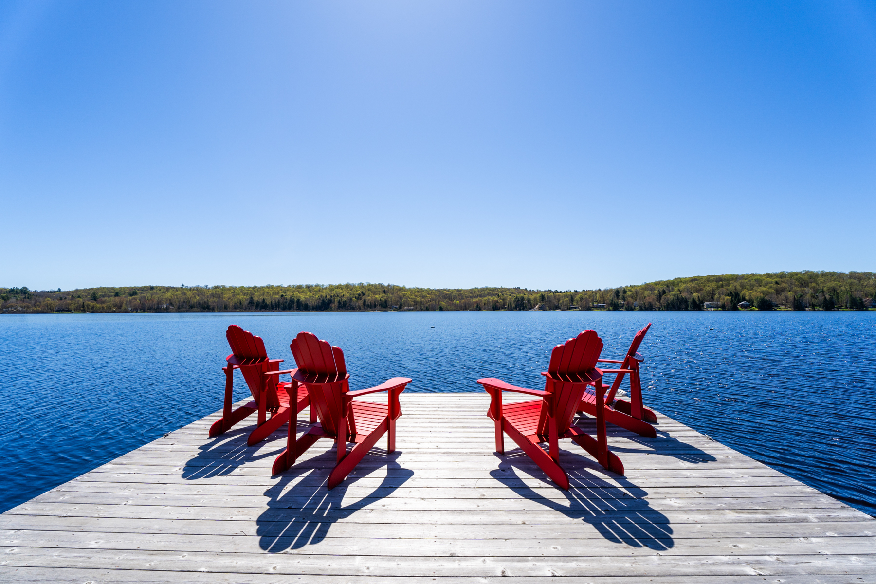 View from the light-coloured dock to the bright blue lake, with four red Muskoka chairs facing the water