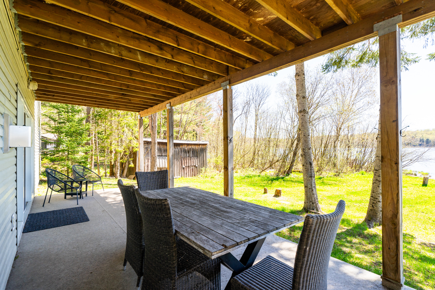 Side angle of a covered lower-level deck with a dining table. In the background, a wood shed