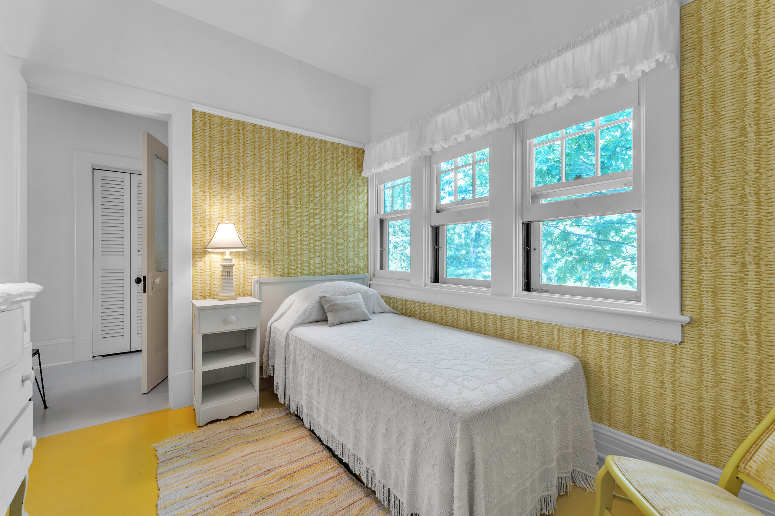 A white twin bed pressed against windows in a yellow room