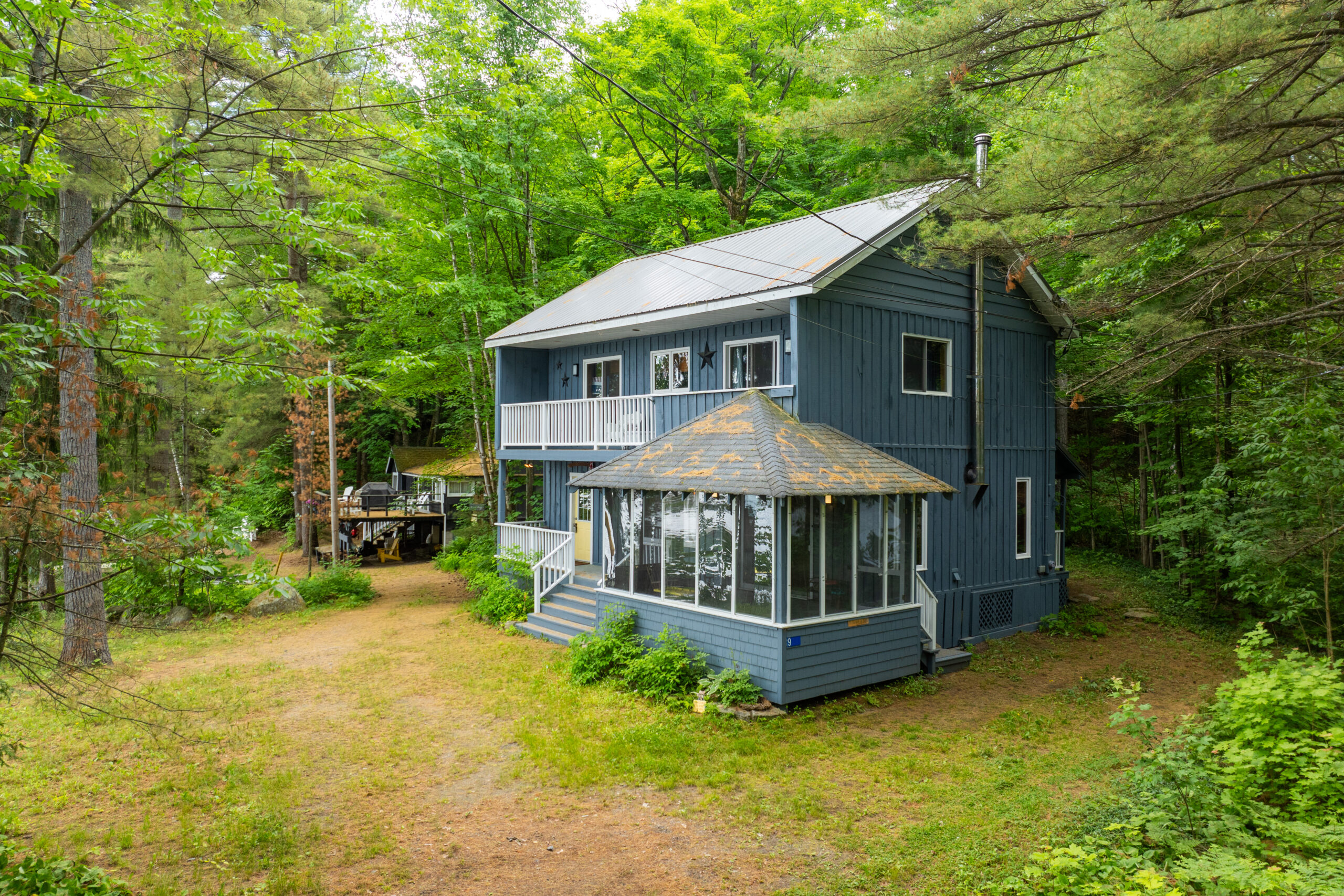 Two-story blue cottage with a large sunroom on the right, surrounded by trees