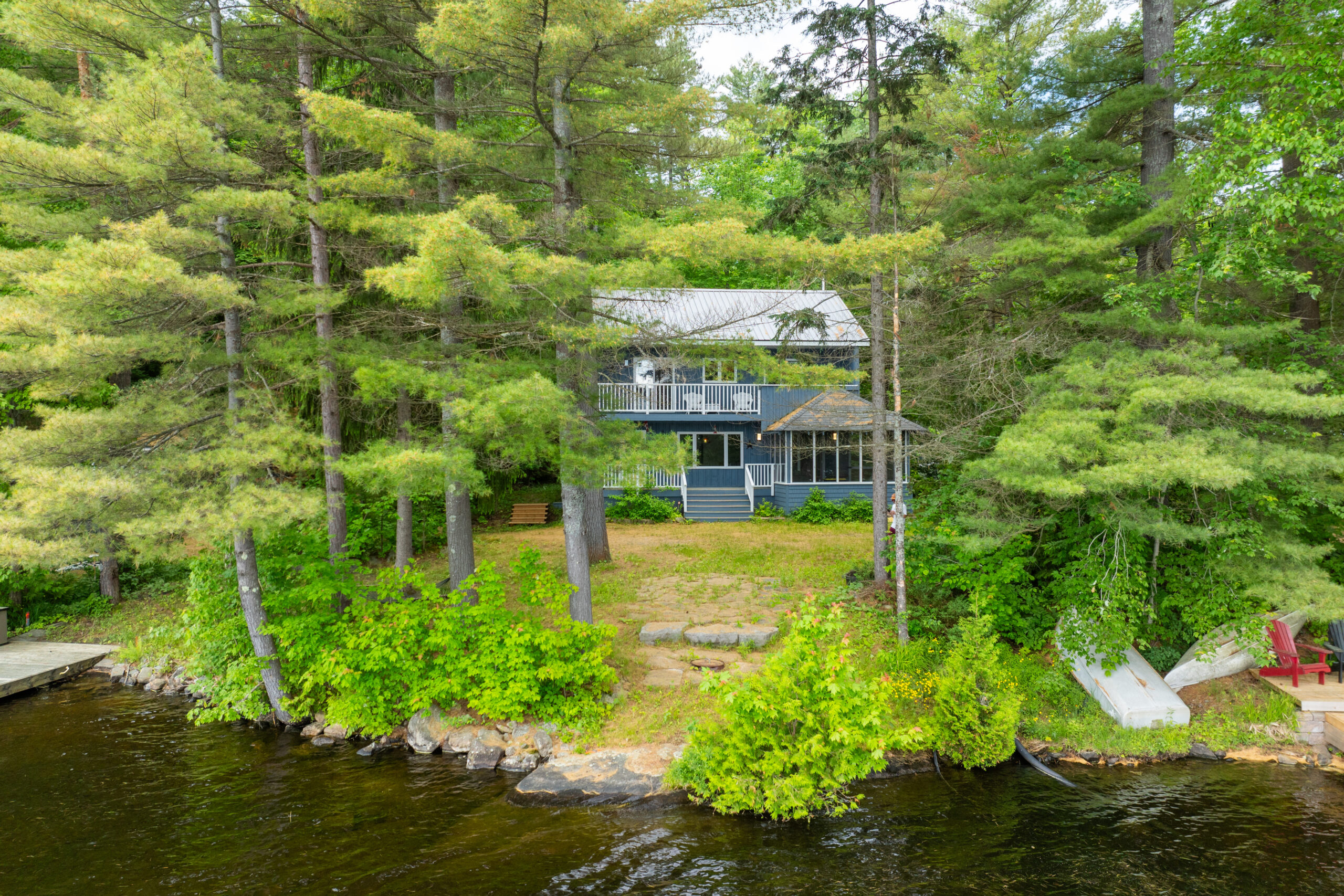 A zoomed out shot of the blue panelled cottage, partially hidden behind bright green trees. A yellow-green grassy yard leads down to the lake