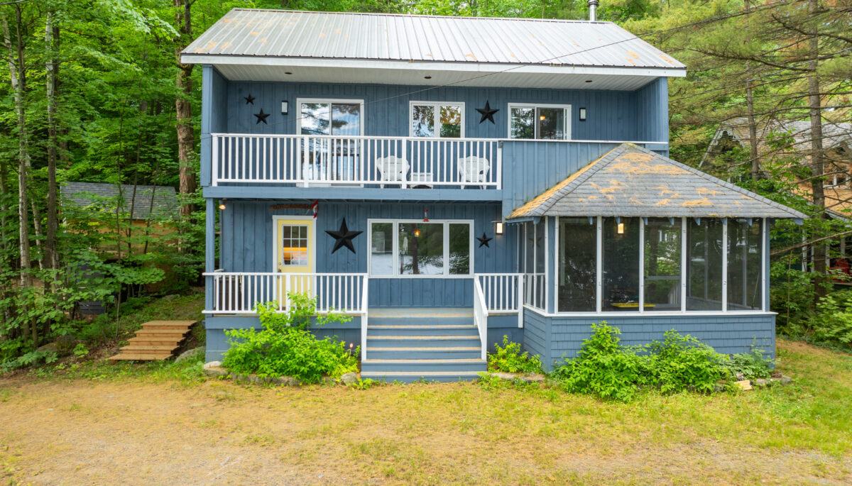 A two-story blue cottage with two balconies and a sunroom attached to the right. On a grassy property