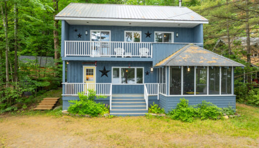 A two-story blue cottage with two balconies and a sunroom attached to the right. On a grassy property