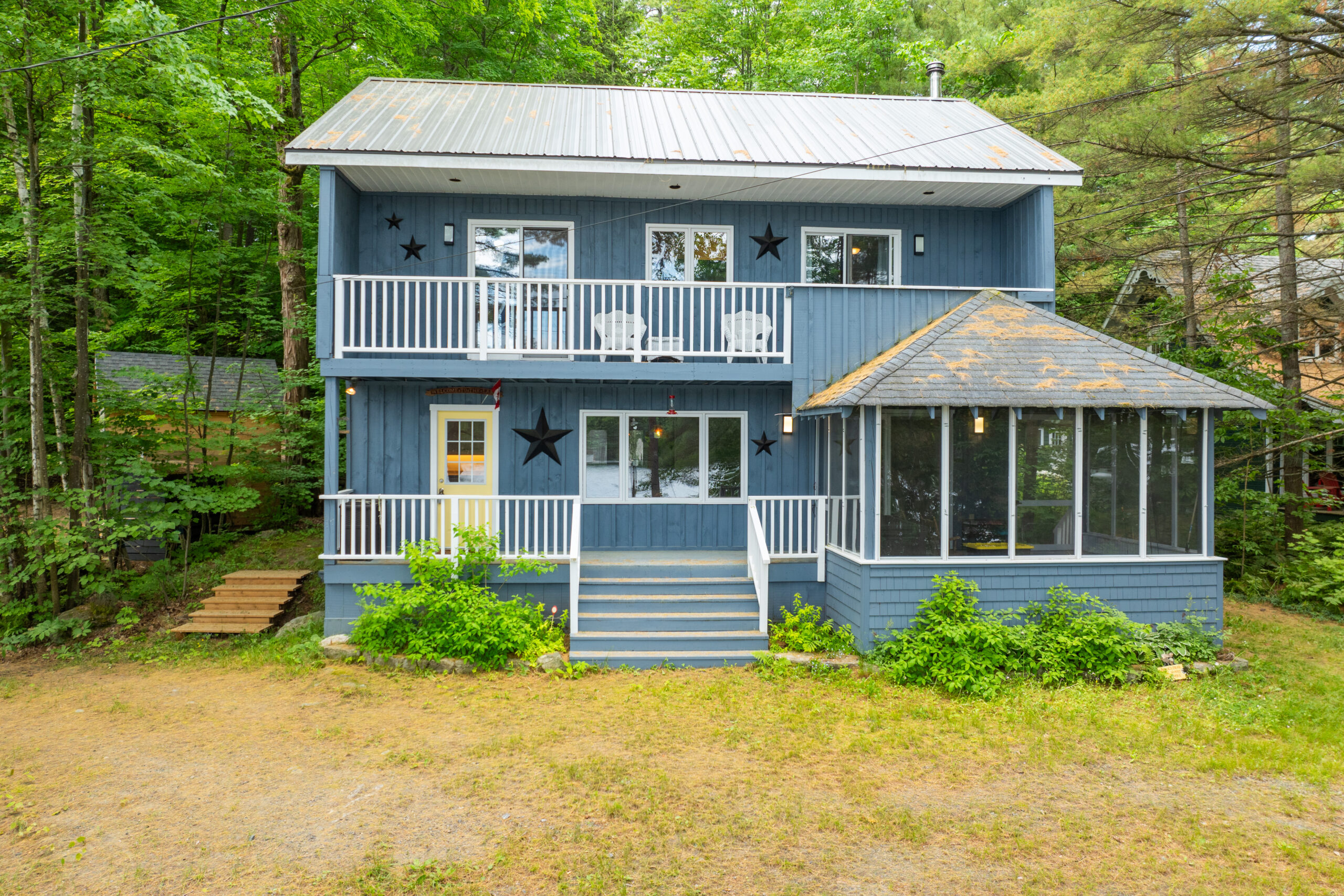 A two-story blue cottage with two balconies and a sunroom attached to the right. On a grassy property