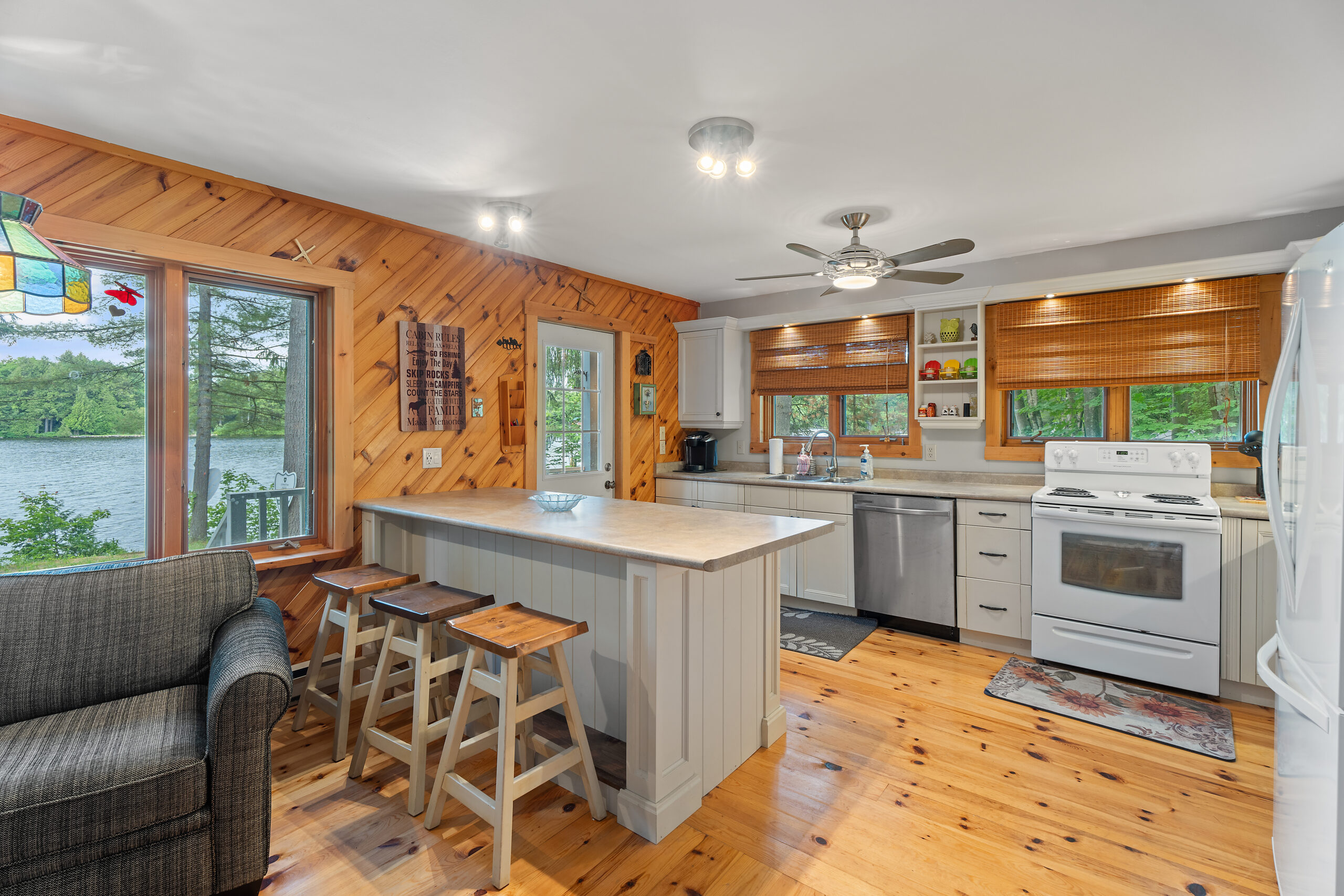 A white kitchen island with barstools faces white cabinets, a stainless steel dishwasher and white oven. The surrounding walls and floors are wood