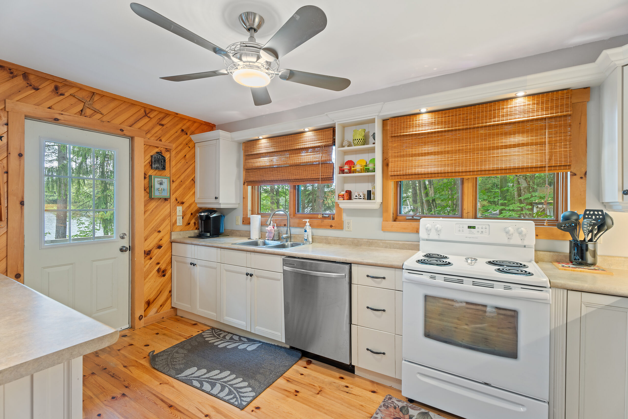 White cabinets, a stainless steel dishwasher, a white oven. Behind, bright windows that look into the forest. A fan hangs overhead