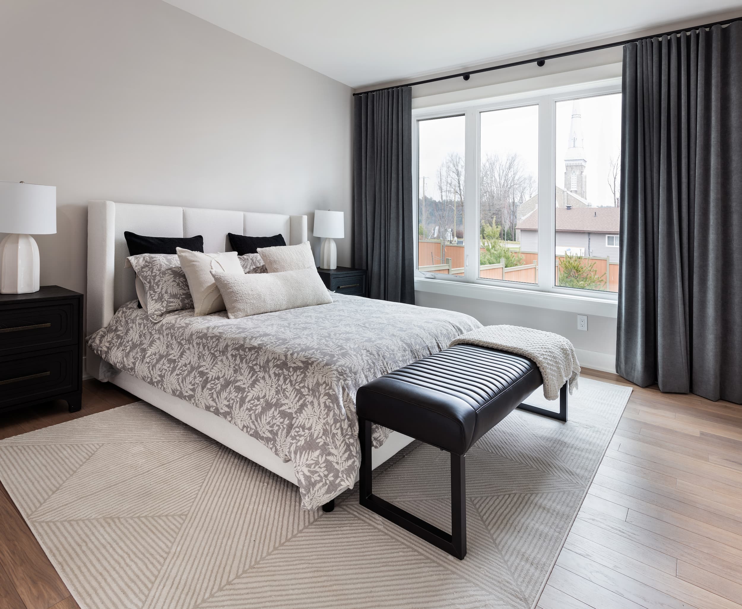 A large bed with a grey cover and a black bench at the end on a cream carpet