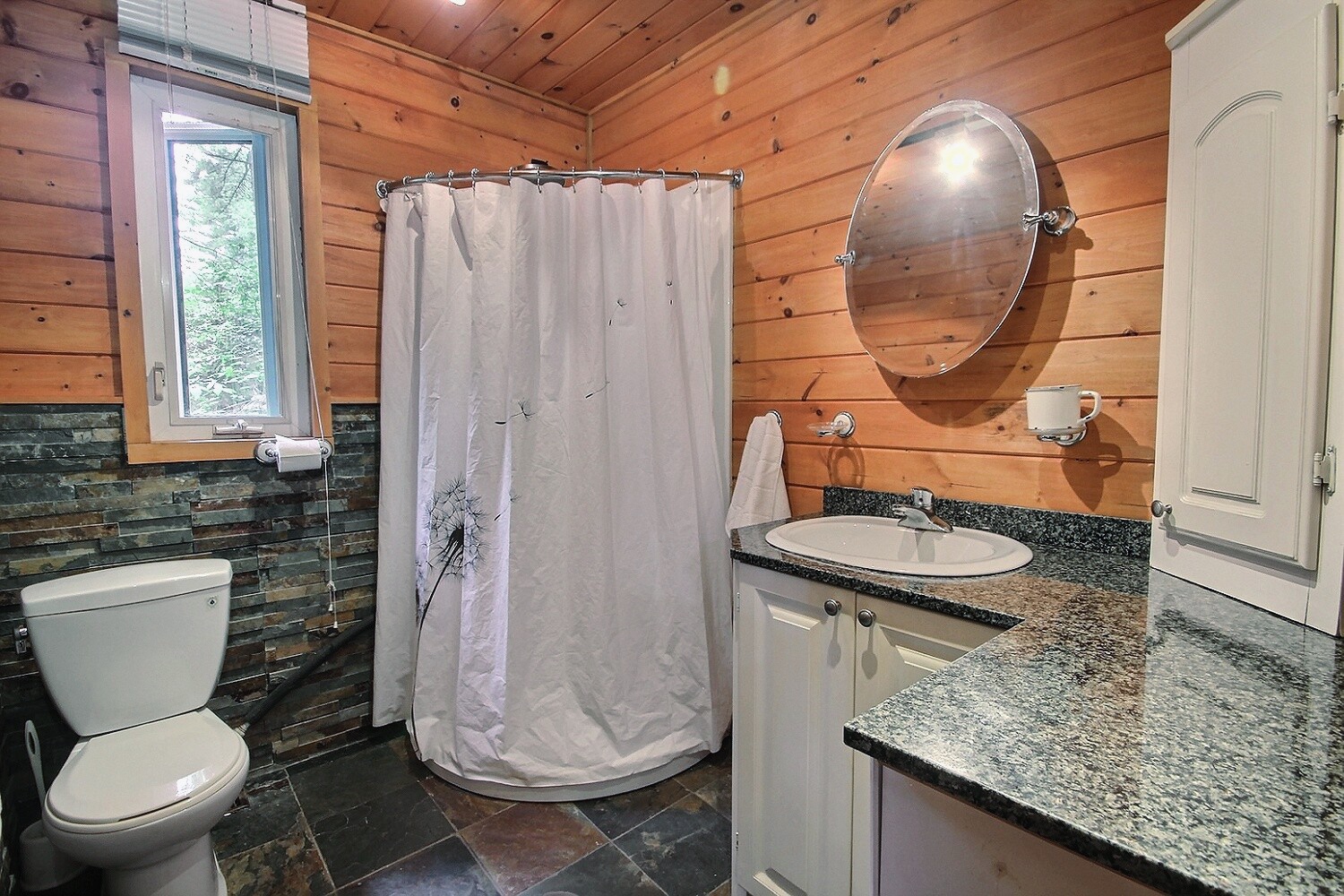 A rounded shower in the corner of the bathroom with a long white shower curtain. To the left, a toilet. To the right, a vanity