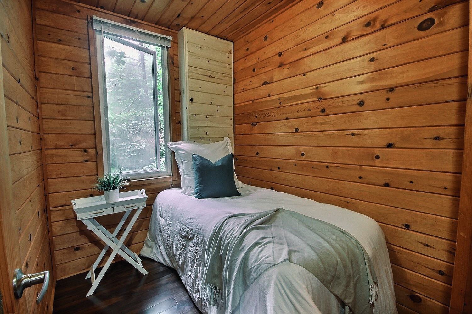 A small white bed with a green blanket pressed against a wall in a wood panelled room