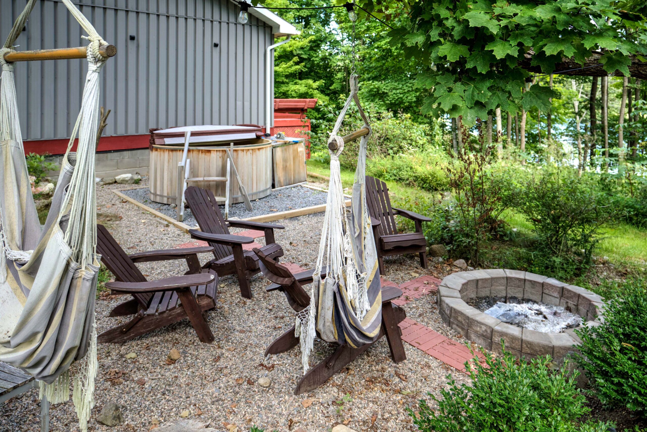 Brown muskoka chairs around a fire pit beside hammocks hanging from a tree