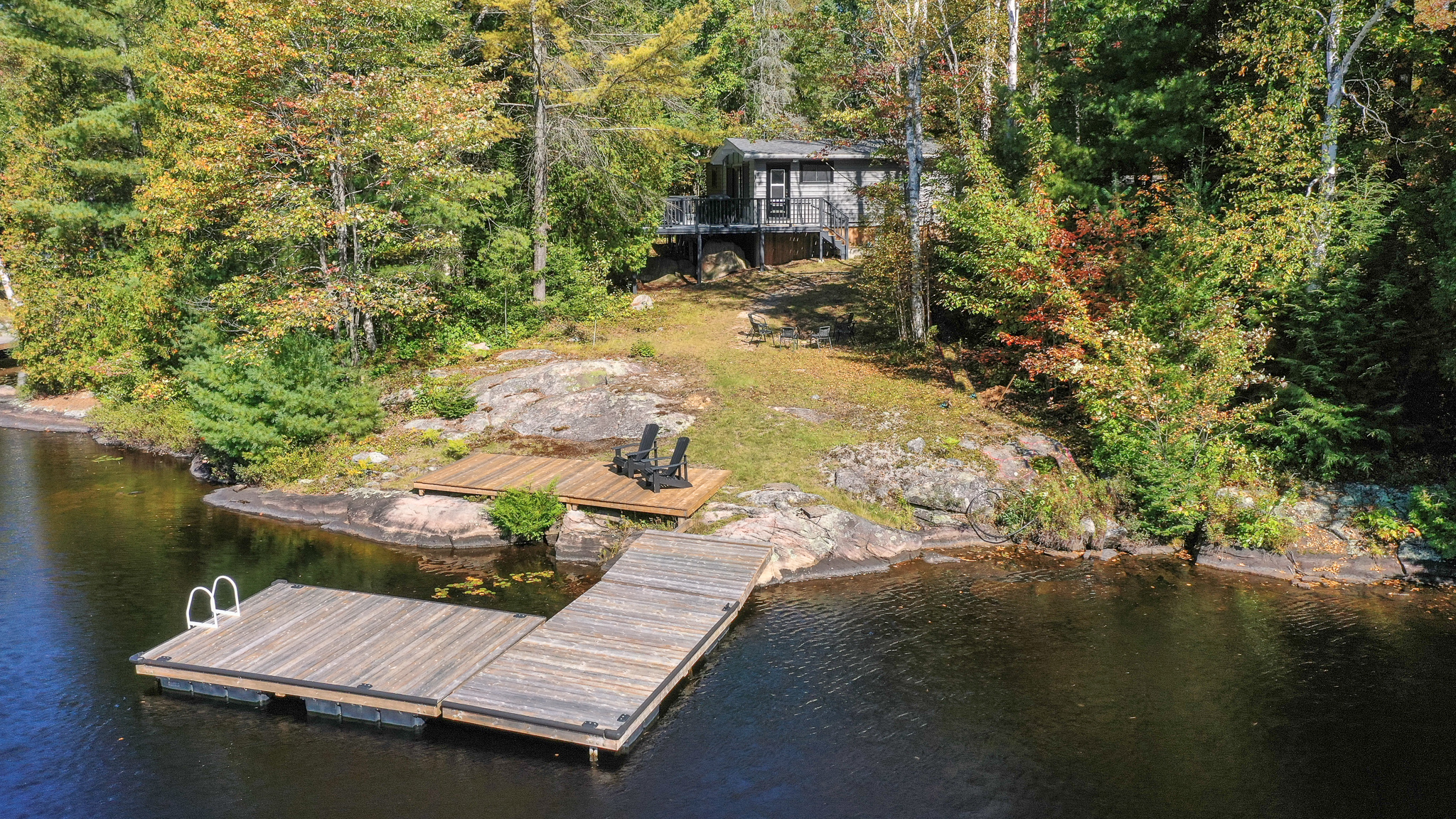 A small cottage is nestled in a forest. An L-shaped dock juts out in the water