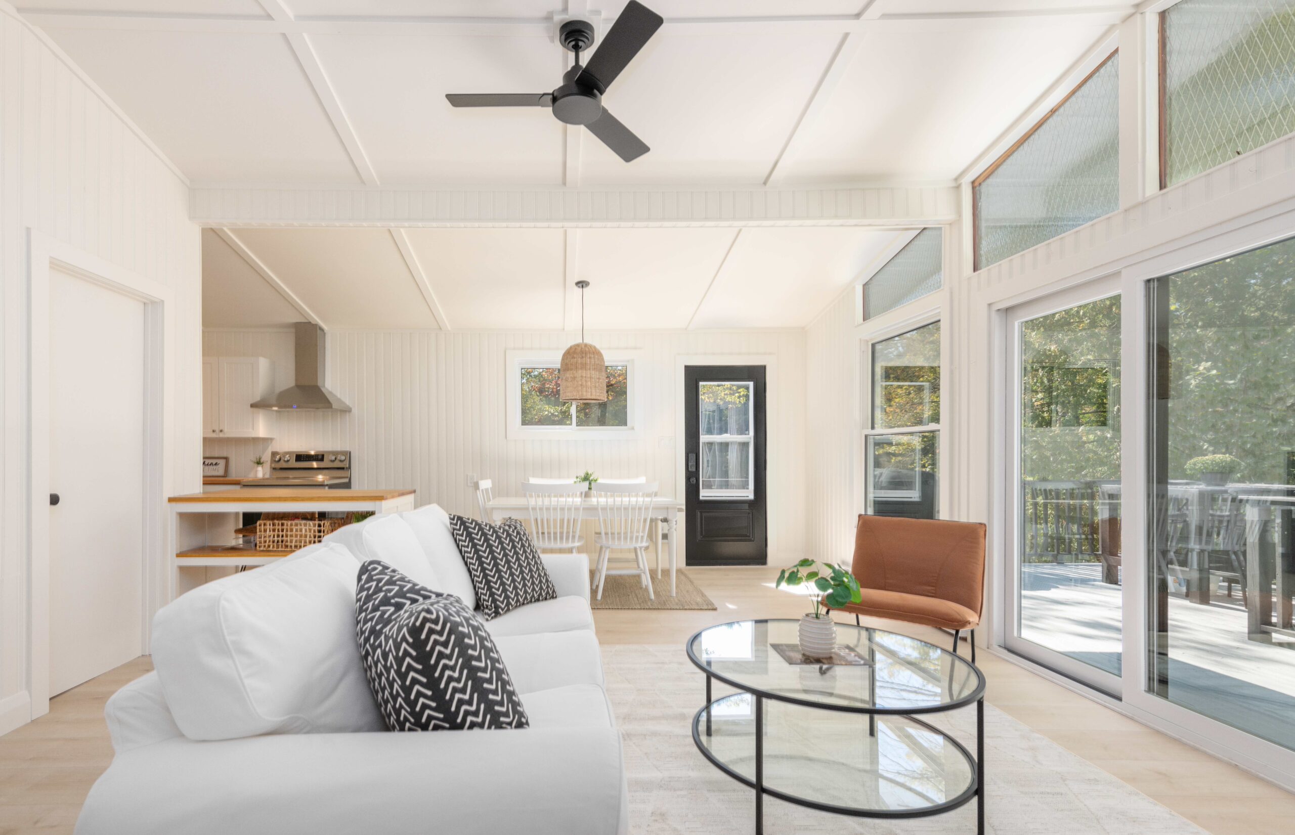 A bright living room with a plush white couch faces a round glass table and a wall of windows