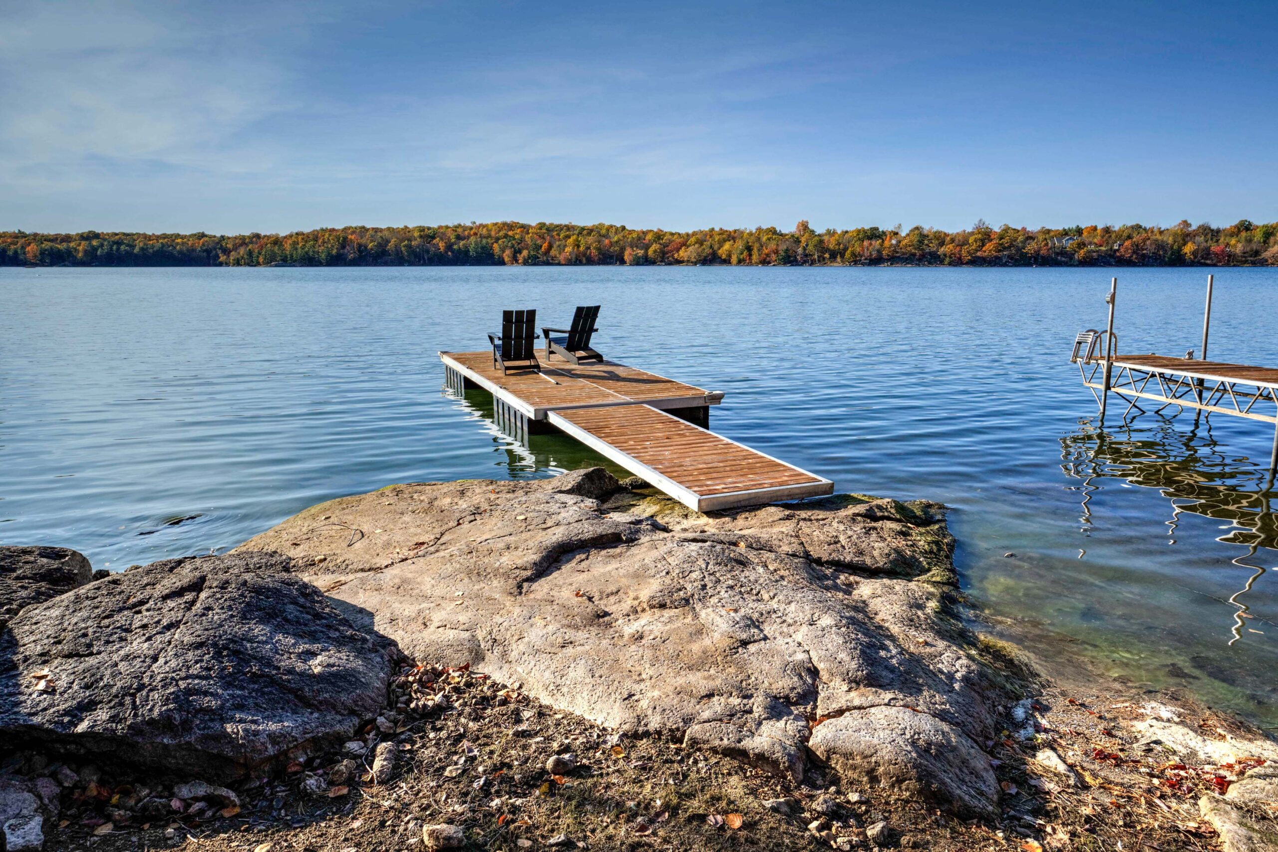 A long brown dock with two Muskoka chairs in blue water