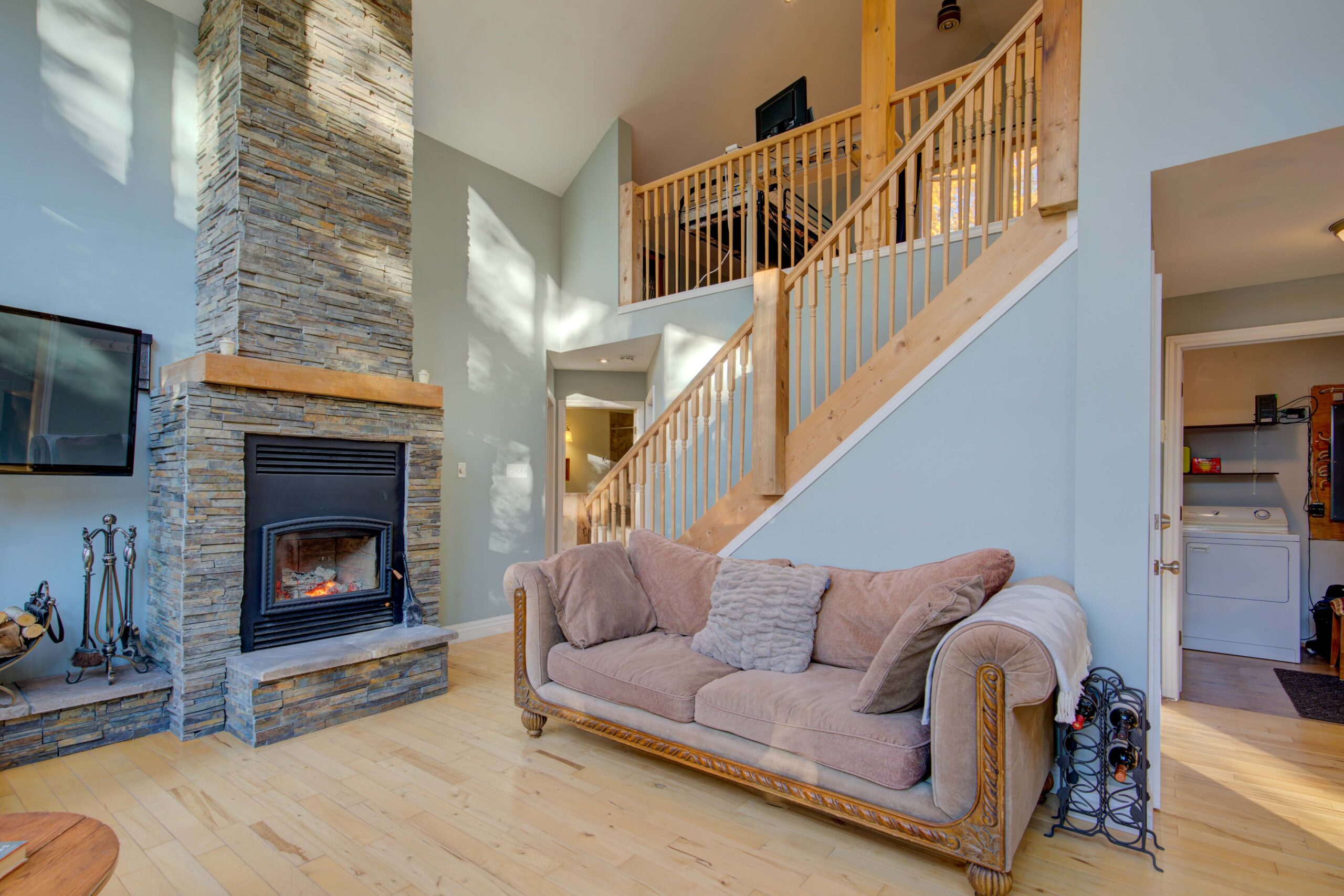 A pink velvet couch sits against a staircase. To the left, a stone fireplace.