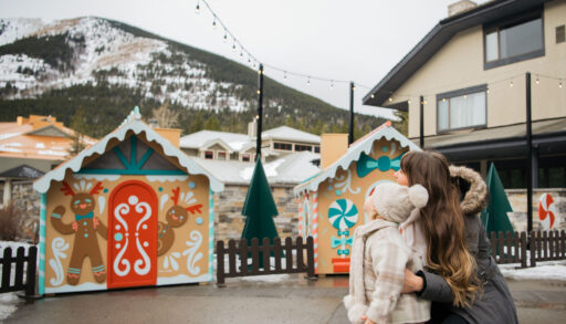 Small gingerbread house shacks stand in front of a large ski kill. A little girl and a woman stare at the shacks.