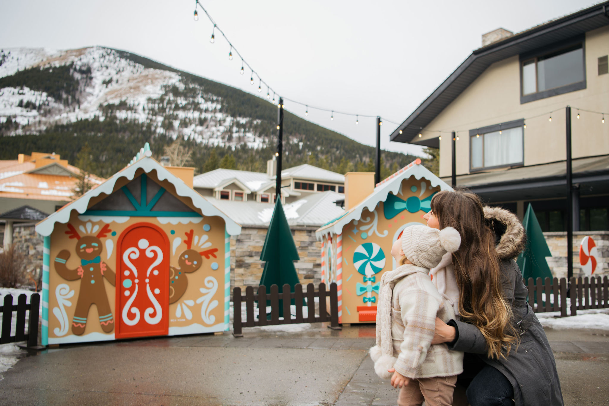 Small gingerbread house shacks stand in front of a large ski kill. A little girl and a woman stare at the shacks.