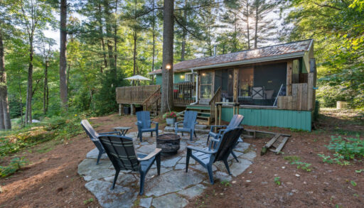 Blue Muskoka chairs sit around a fire pit on a stone area beside the teal cottage