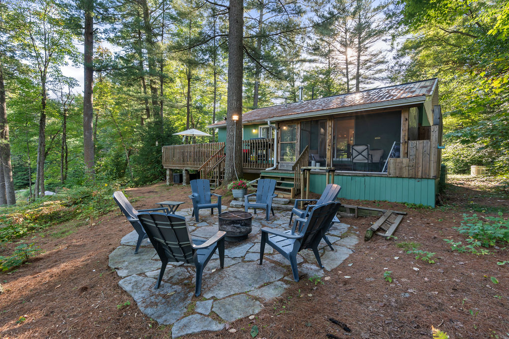 Blue Muskoka chairs sit around a fire pit on a stone area beside the teal cottage