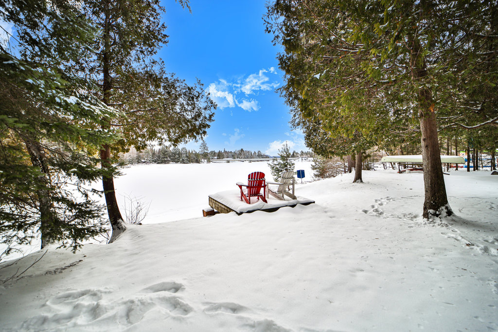 A snowy lakeside with two red Muskoka chairs facing the snowed-over lake