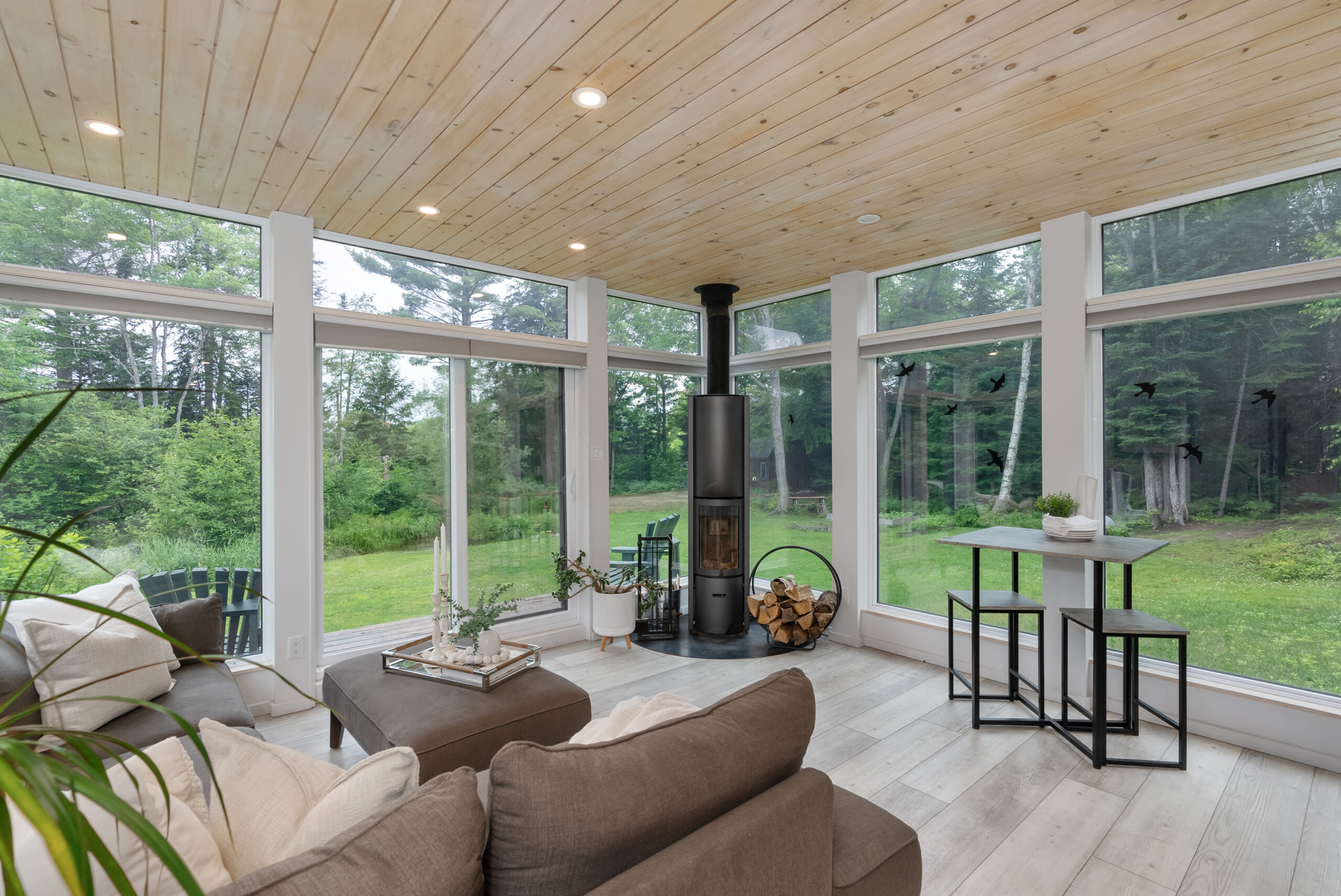 A grey couch faces a slim wood fireplace in a glass room with wood ceilings