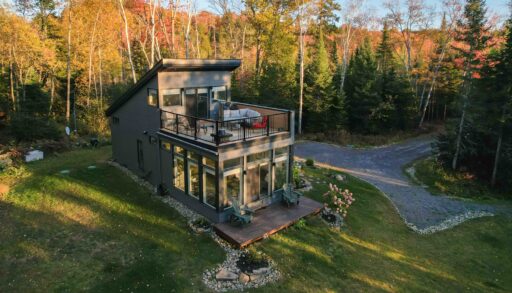 A dark grey cottage with a second-story deck on a large grassy property