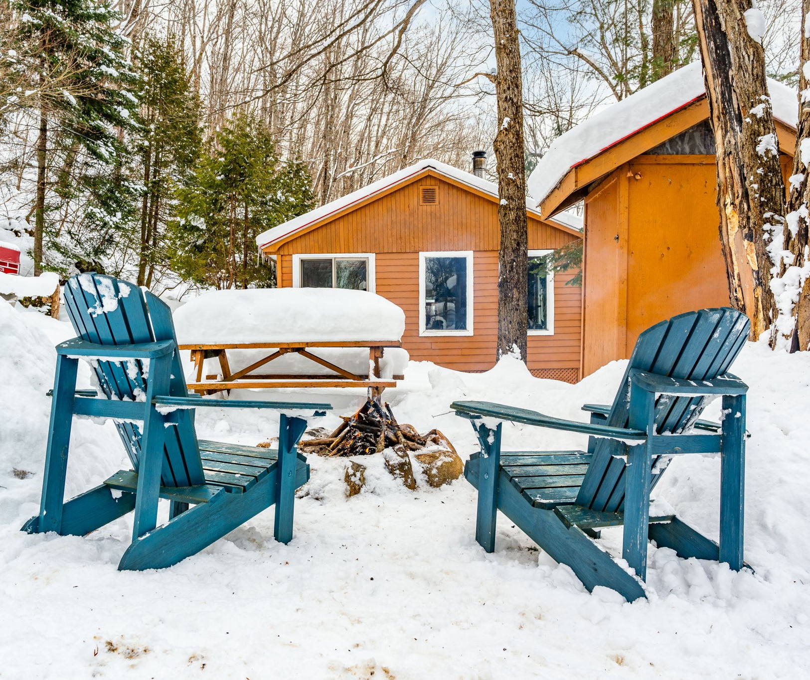 Two turquoise Muskoka chairs face a fire pit. Behind, a wood outhouse and a wood cabin