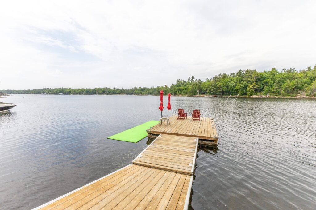 A long wood dock with two red Muskoka chairs at the end. A bright green floating lily pad sits in the water beside it