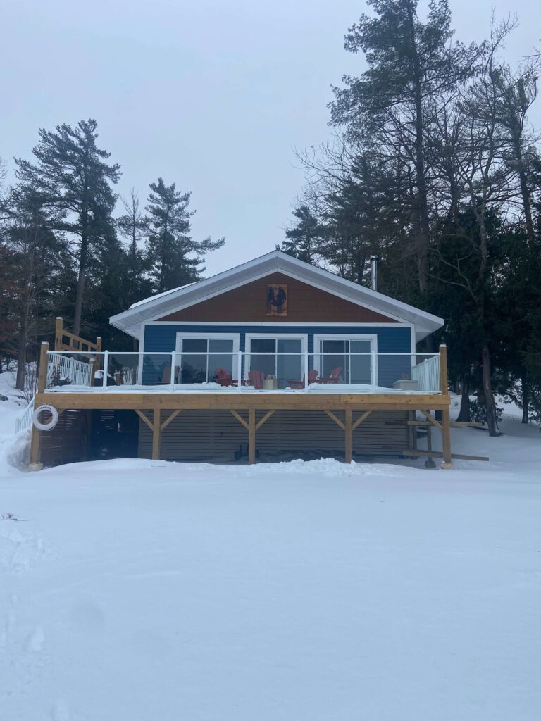 A blue-paneled cottage with a glass-framed deck on a snowy lawn. Three red Muskoka chairs sit in front